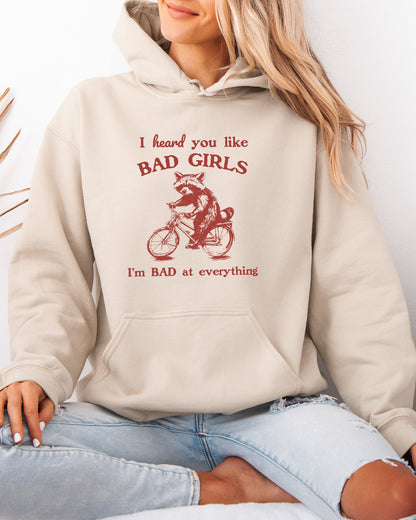 Woman wearing a sand beige hoodie featuring a funny retro-style raccoon riding a bycicle graphic and the quote 'I Heard You Like Bad Girls, I Am Bad At Everything'.