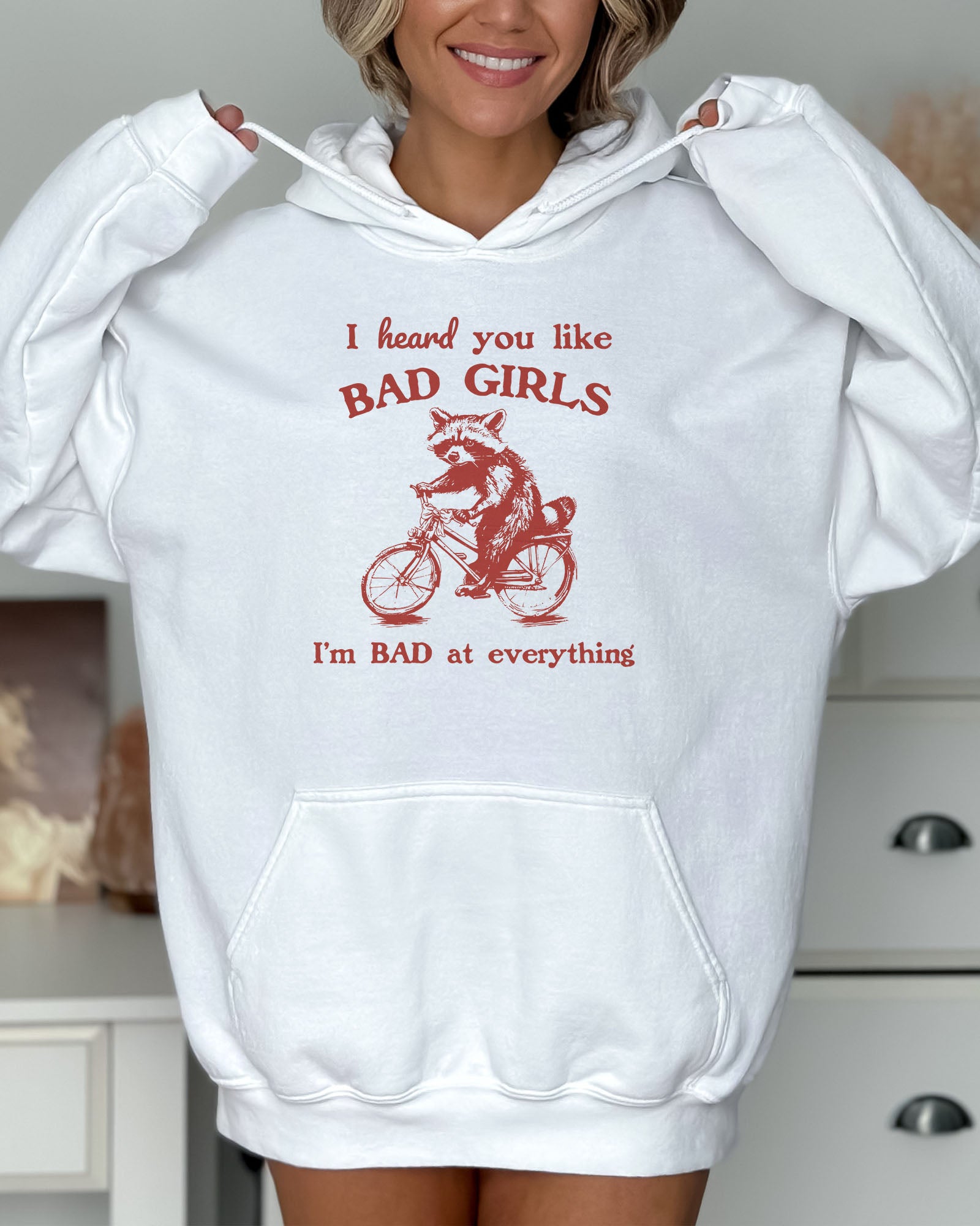 Woman wearing a white hoodie featuring a funny retro-style raccoon riding a bycicle graphic and the quote 'I Heard You Like Bad Girls, I Am Bad At Everything'.