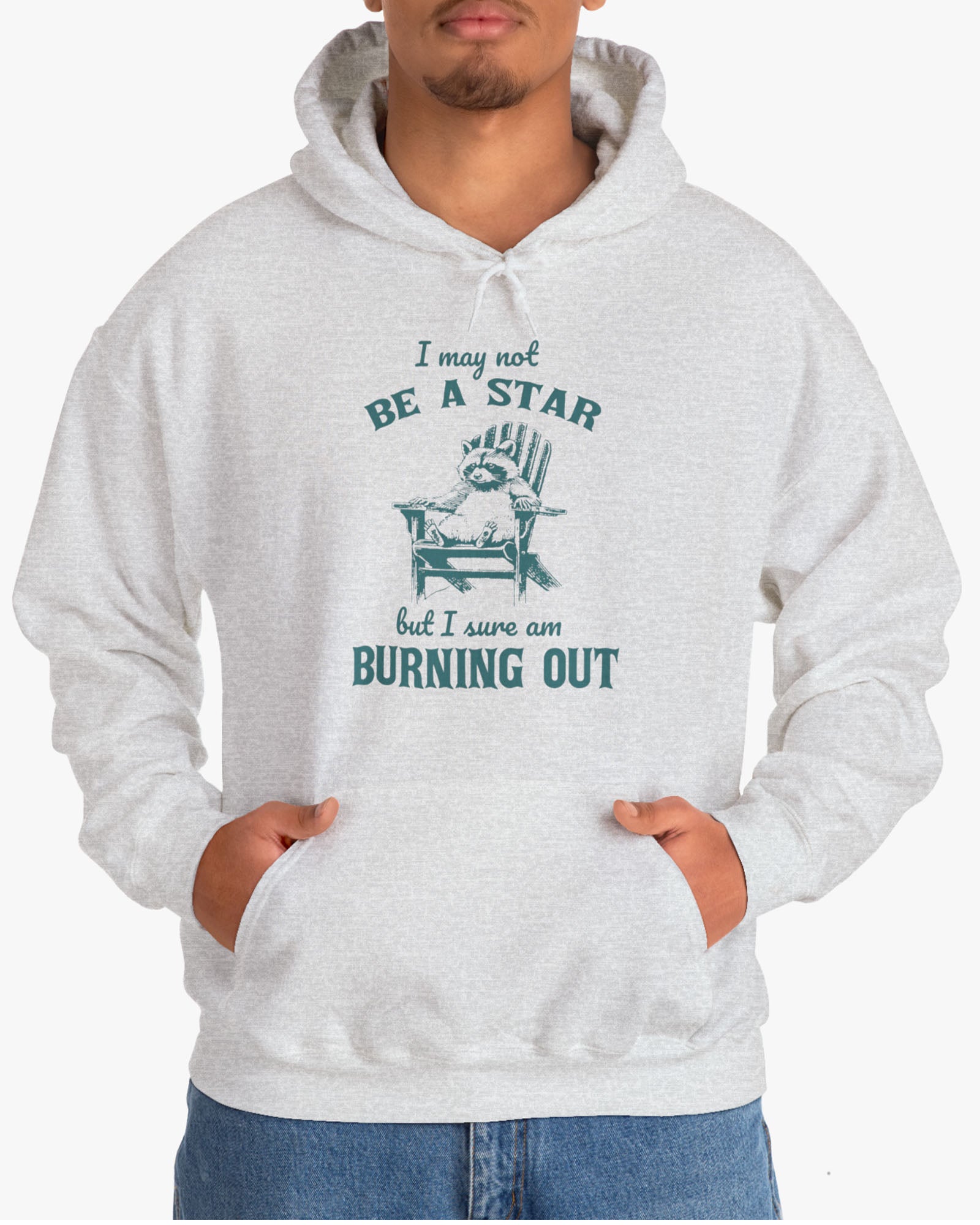 Man wearing a 'I May Not Be A Star But I Sure Am Burning Out' hoodie in ash gray, featuring a retro raccoon graphic design and unisex fit.