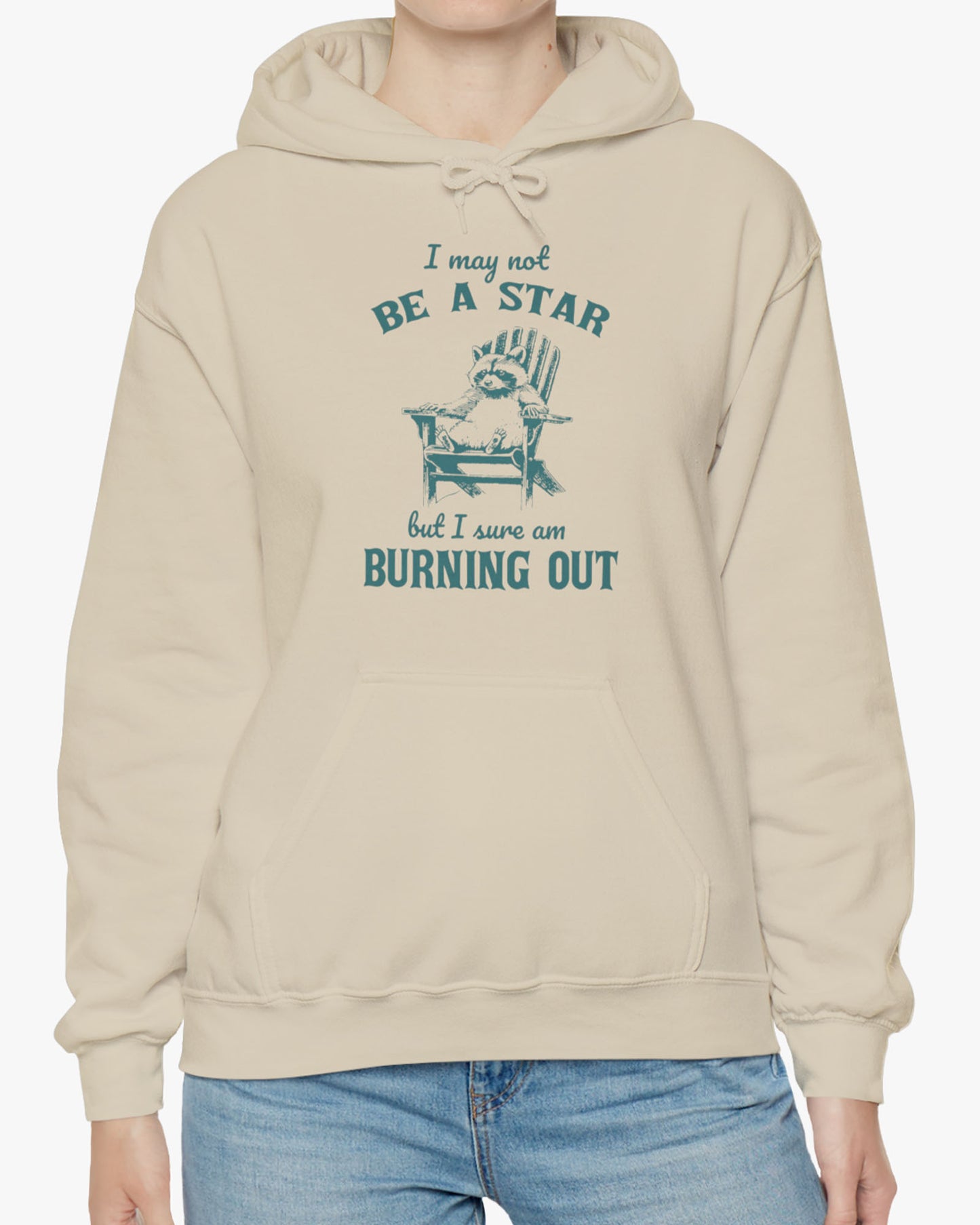 Woman wearing a 'I May Not Be A Star But I Sure Am Burning Out' hoodie in sandy beige, featuring a retro raccoon graphic design and unisex fit.