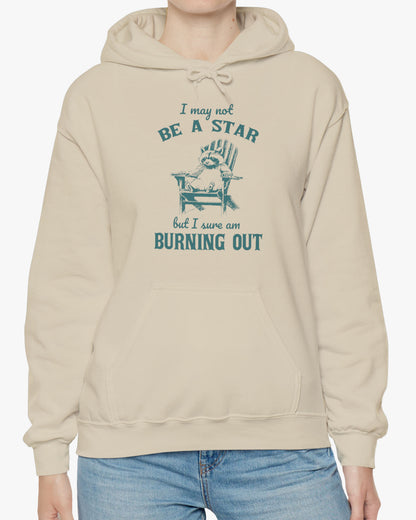 Woman wearing a 'I May Not Be A Star But I Sure Am Burning Out' hoodie in sandy beige, featuring a retro raccoon graphic design and unisex fit.