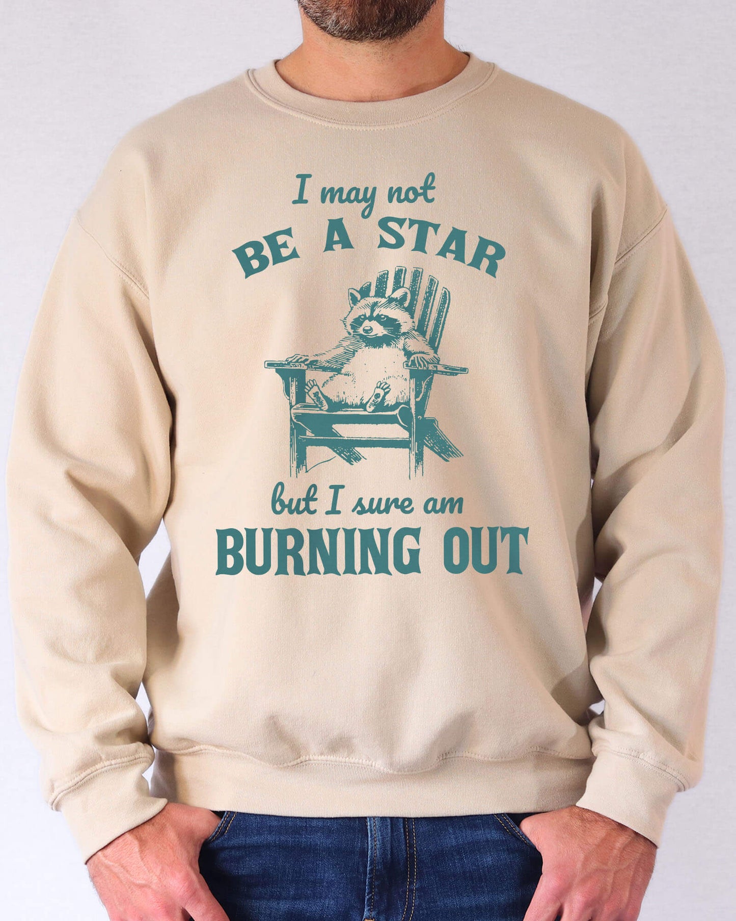 Man wearing a sand beige sweatshirt featuring a funny retro graphic of a raccoon sitting on a chair and the quote 'I May Not Be A Star But I Sure Am Burning Out'.