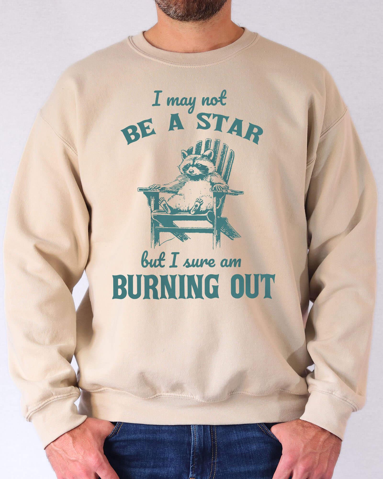 Man wearing a sand beige sweatshirt featuring a funny retro graphic of a raccoon sitting on a chair and the quote 'I May Not Be A Star But I Sure Am Burning Out'.