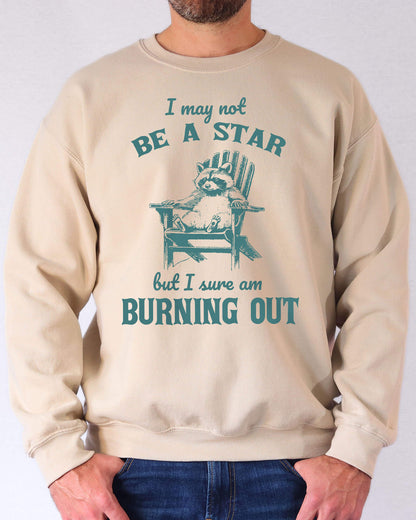 Man wearing a sand beige sweatshirt featuring a funny retro graphic of a raccoon sitting on a chair and the quote 'I May Not Be A Star But I Sure Am Burning Out'.
