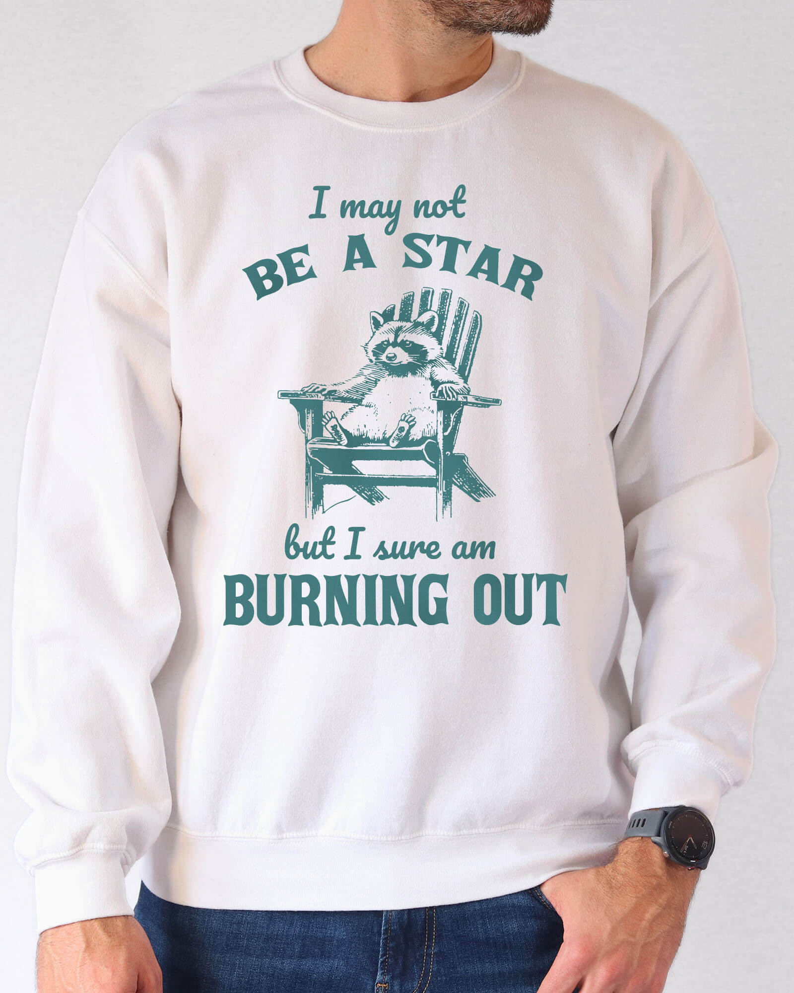 Man wearing a white sweatshirt featuring a funny retro graphic of a raccoon sitting on a chair and the quote 'I May Not Be A Star But I Sure Am Burning Out'.