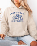 Woman wearing a sand beige hoodie featuring a funny retro-style raccoon gang graphic and the quote 'I Put The Sexy In Dyslexia'.