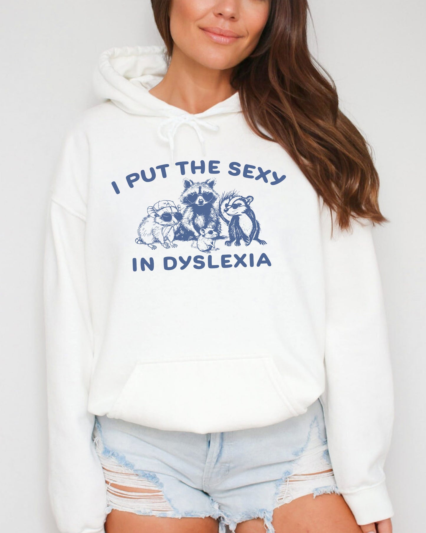 Woman wearing a white hoodie featuring a funny retro-style raccoon gang graphic and the quote 'I Put The Sexy In Dyslexia'.