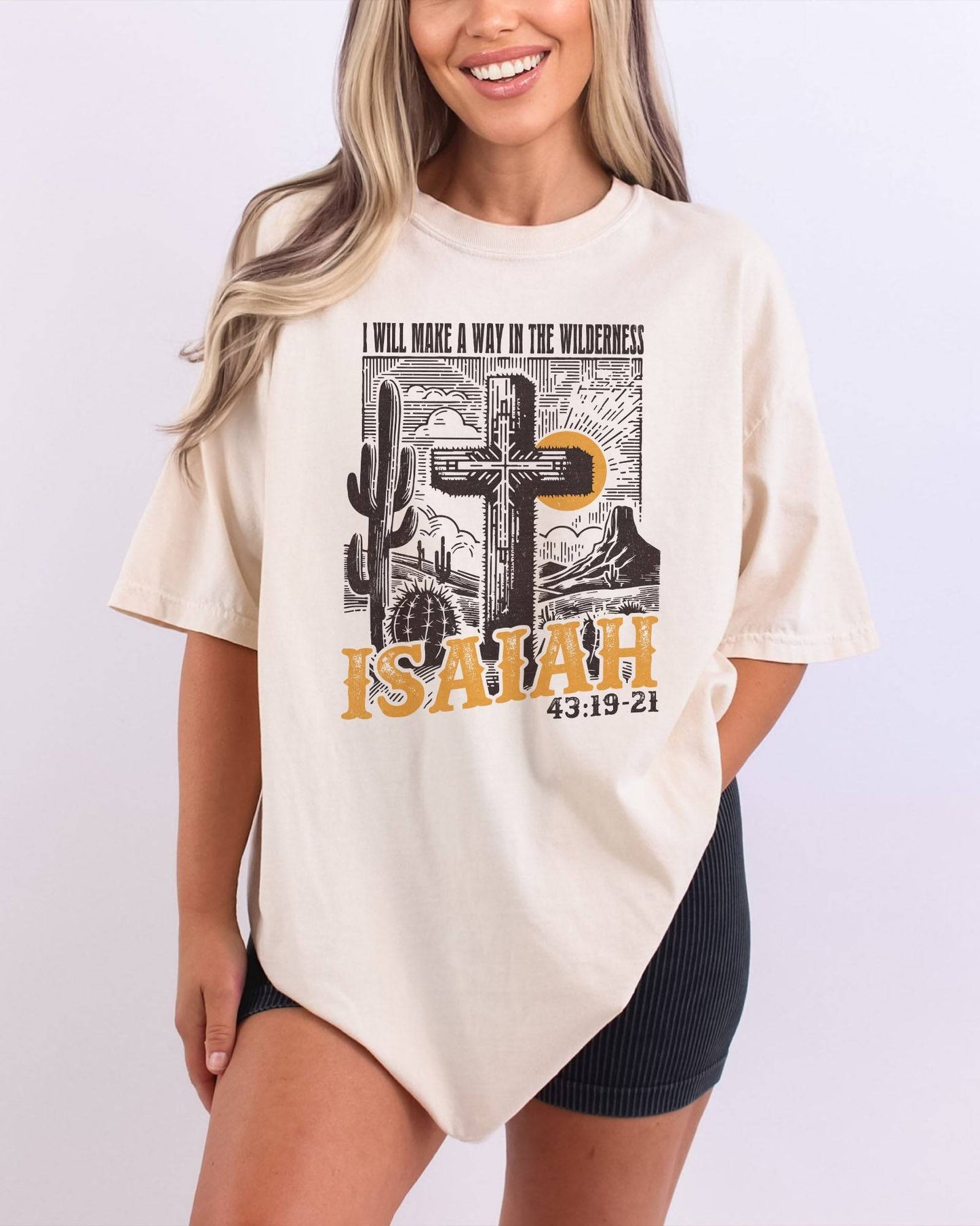 Woman wearing an ivory 'I Will Make a Way in the Wilderness – Isaiah 43:19-21' shirt with a retro graphic of a western desert and a Christian cross.