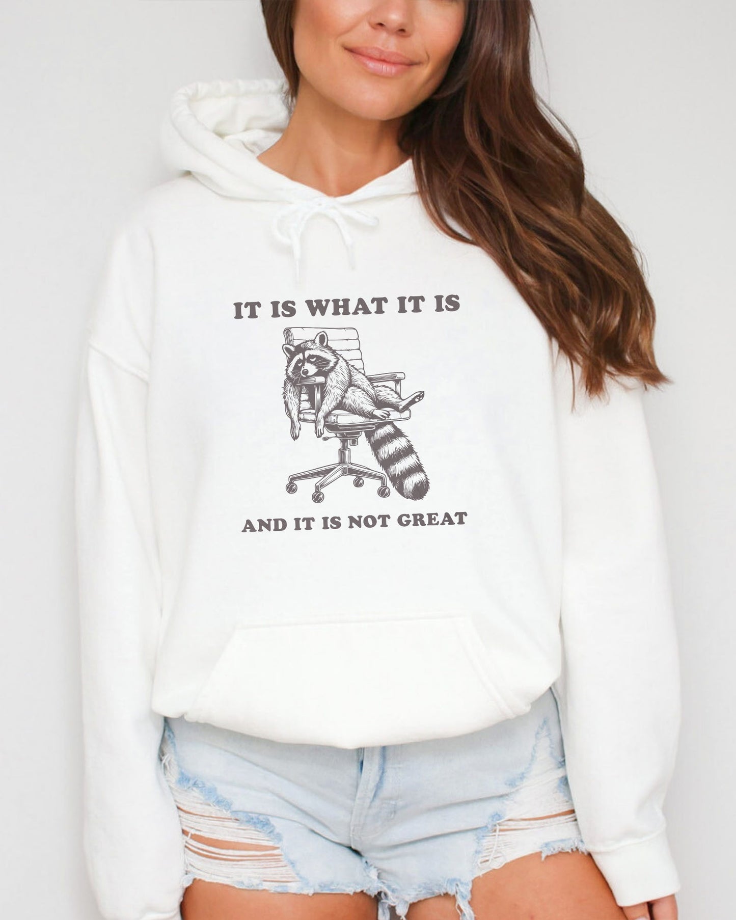 Woman wearing a white 'It Is What It Is And It Is Not Great' hoodie with a funny retro graphic of an exhausted raccoon sitting on a chair.