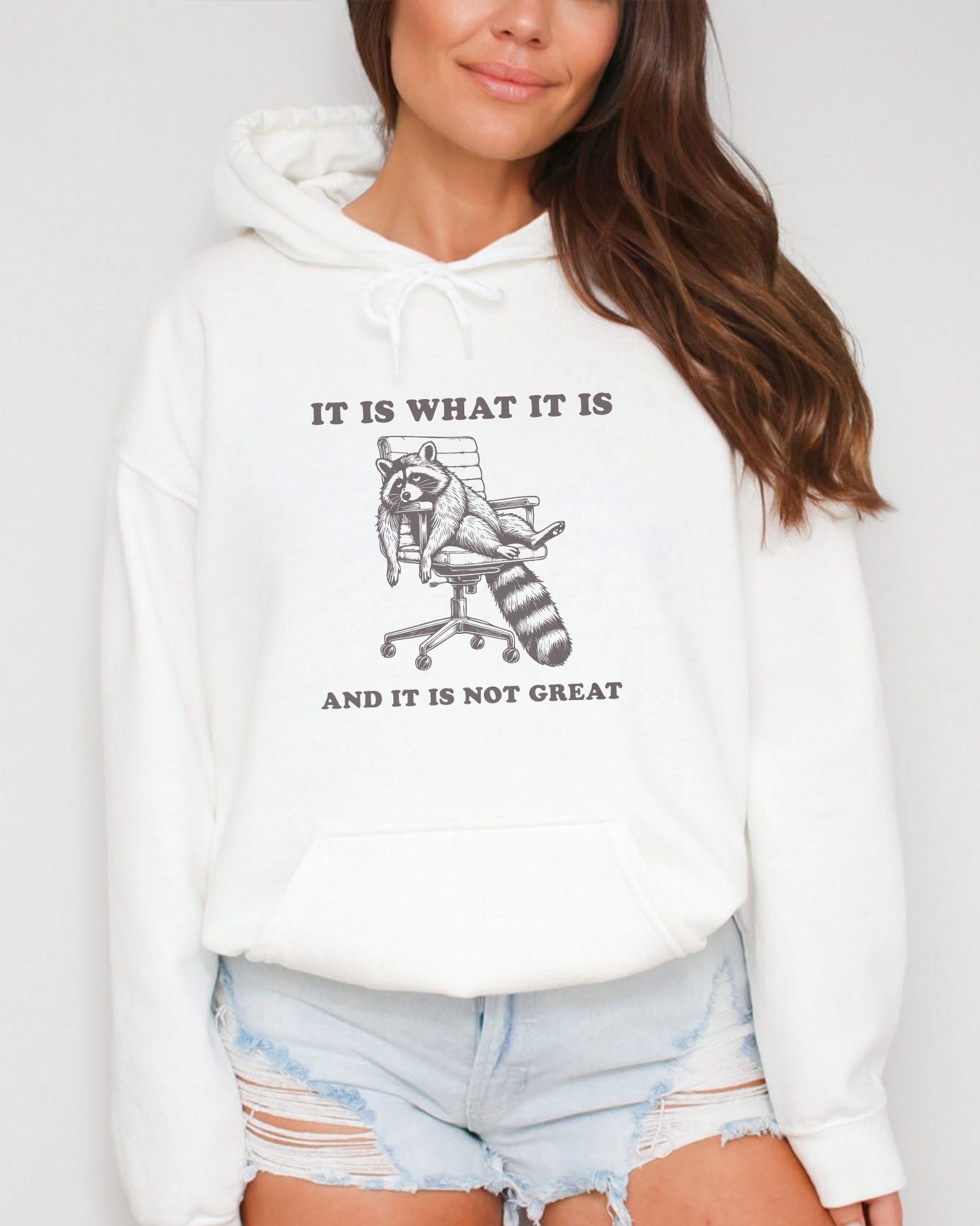 Woman wearing a white 'It Is What It Is And It Is Not Great' hoodie with a funny retro graphic of an exhausted raccoon sitting on a chair.