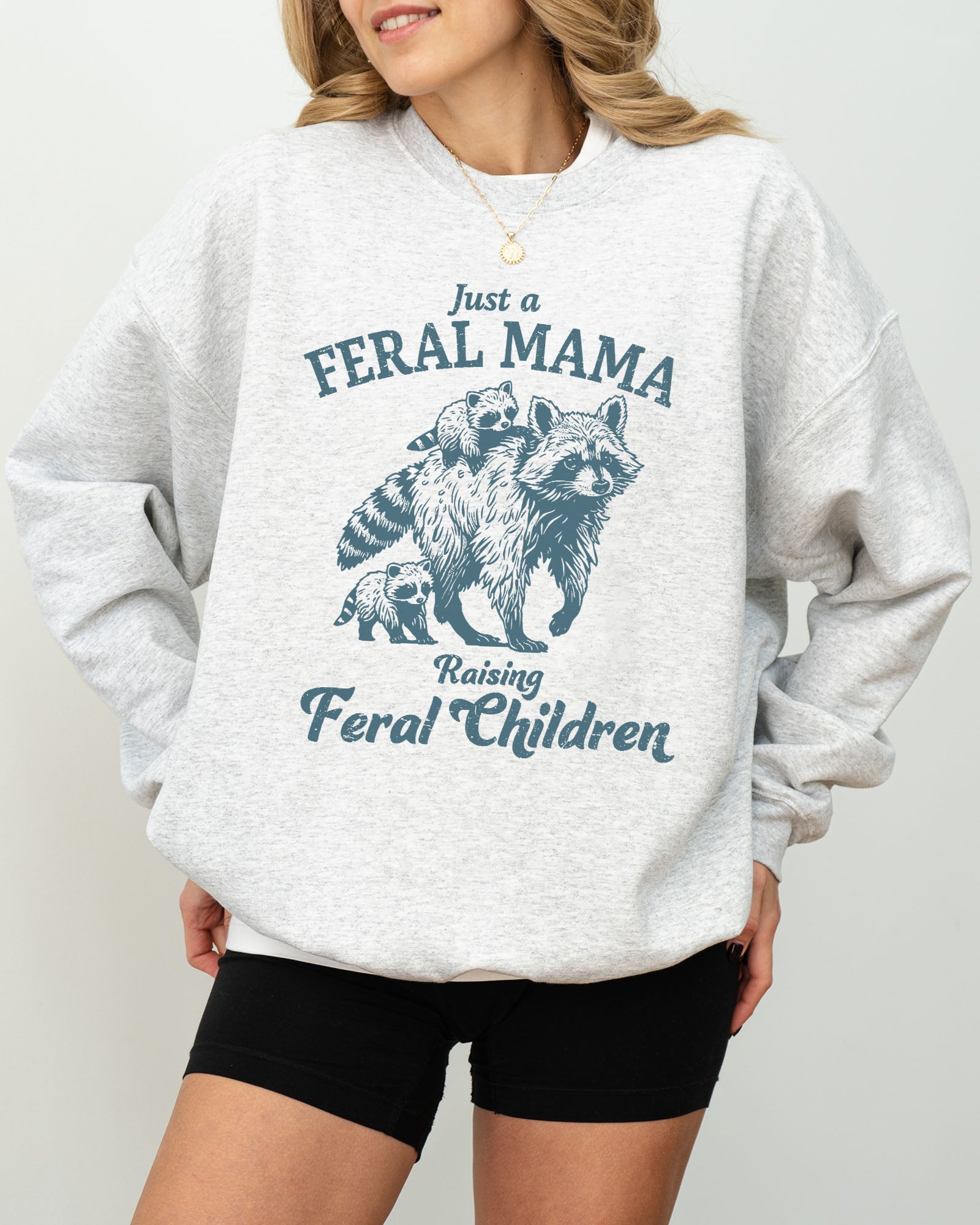 Model wearing ash color sweatshirt with 'Just A Feral Mama Raising Feral Children' raccoons graphic design