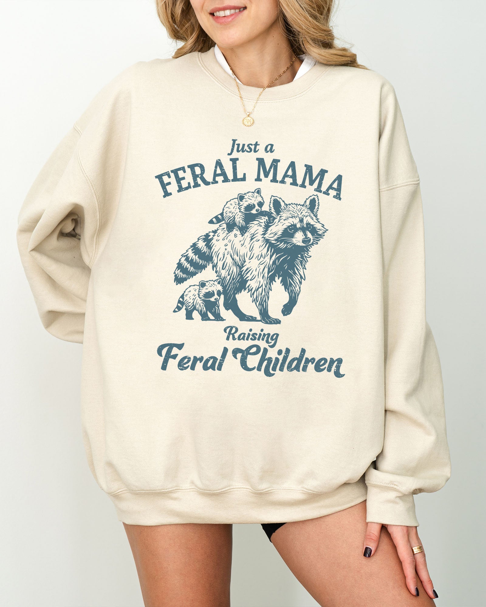 Model wearing sand color sweatshirt with 'Just A Feral Mama Raising Feral Children' raccoons graphic design
