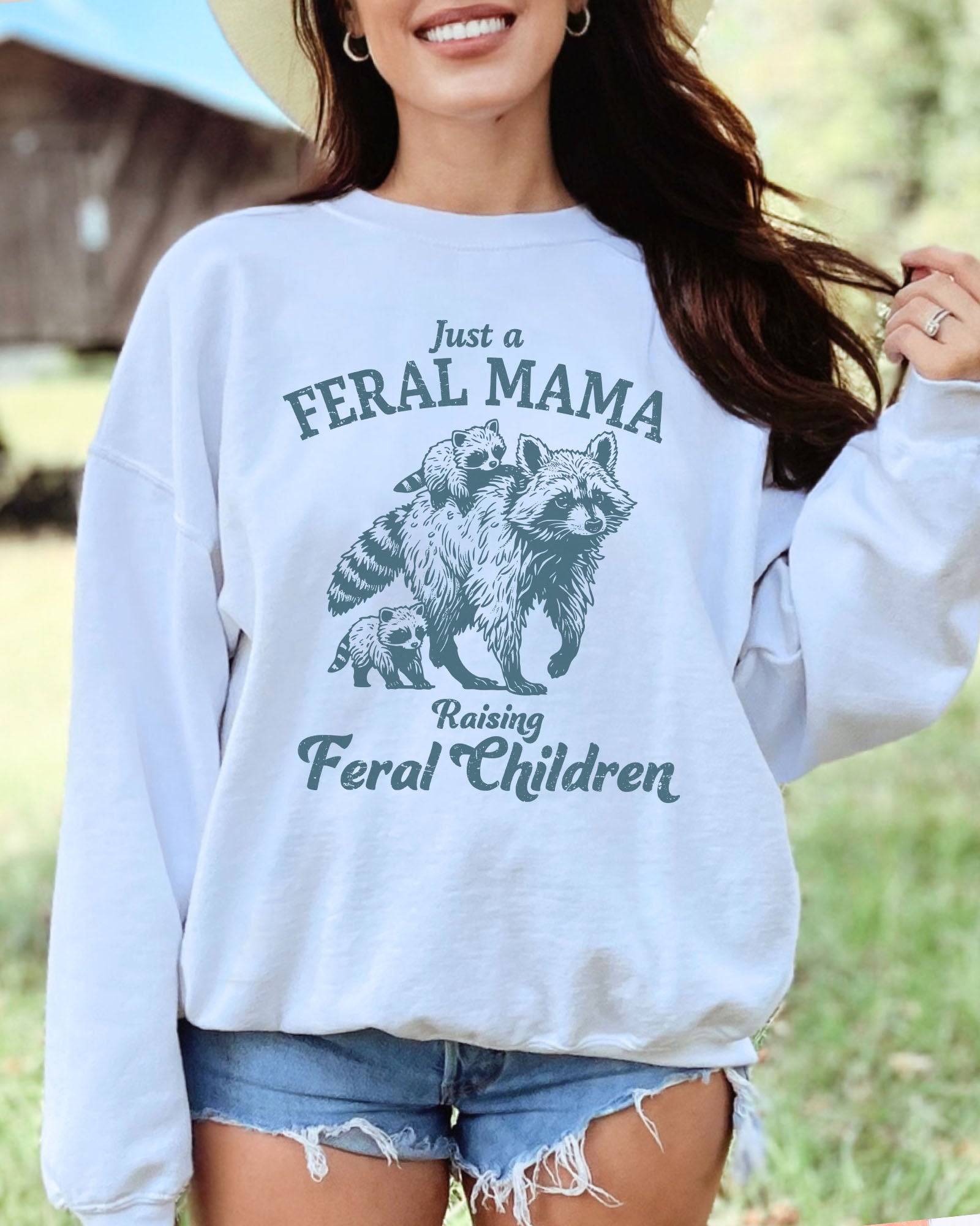 Model wearing white color sweatshirt with 'Just A Feral Mama Raising Feral Children' raccoons graphic design