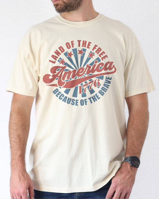 Model wearing natural color shirt with 'Land Of The Free Because Of The Brave' graphic design