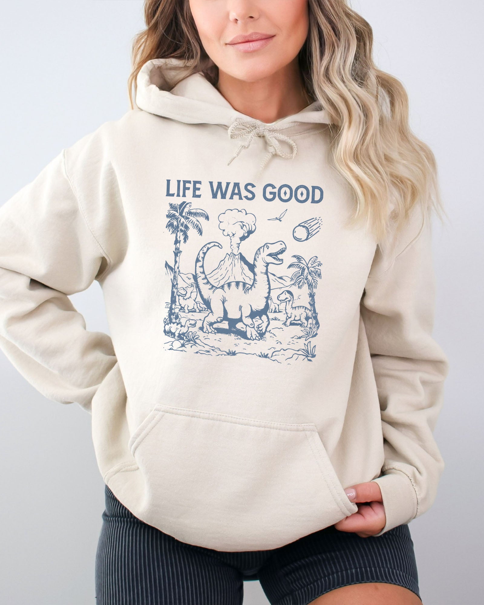 Woman wearing a sand beige hoodie featuring a retro-style graphic of dinosaurs going extinct from an asteroid, with the text 'Life Was Good'.