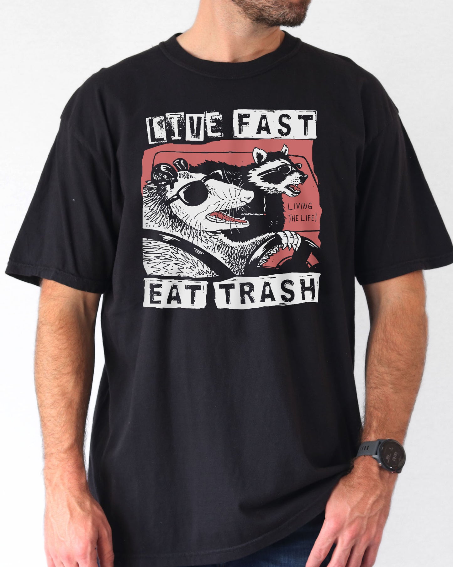 Man  wearing a black-colored "Live Fast Eat Trash" shirt with a retro graphic of a raccoon and opossum driving a car.