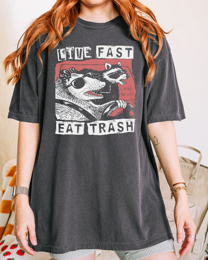 Woman wearing a pepper-colored "Live Fast Eat Trash" shirt with a retro graphic of a raccoon and opossum driving a car.