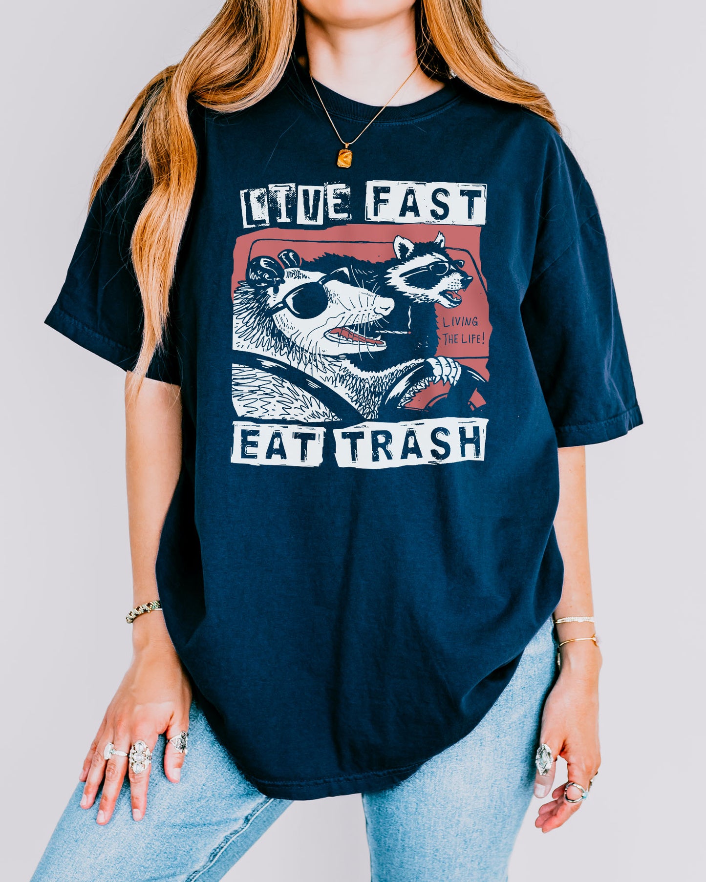Woman wearing a true-navy-colored "Live Fast Eat Trash" shirt with a retro graphic of a raccoon and opossum driving a car.