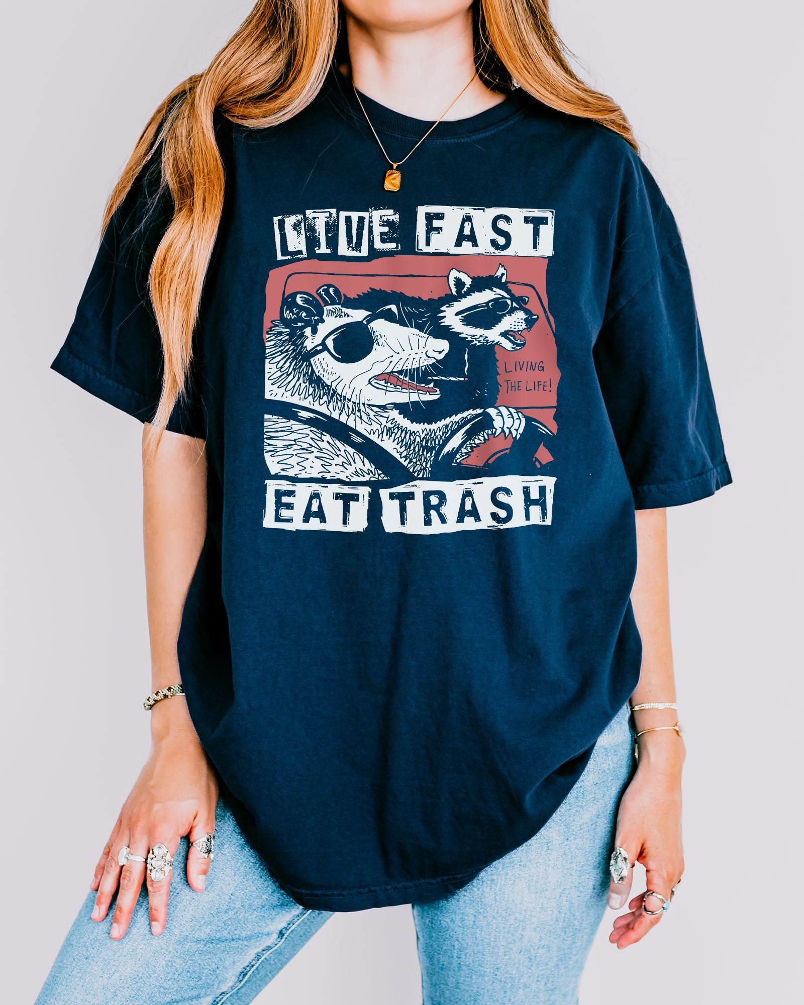 Woman wearing a true-navy-colored "Live Fast Eat Trash" shirt with a retro graphic of a raccoon and opossum driving a car.