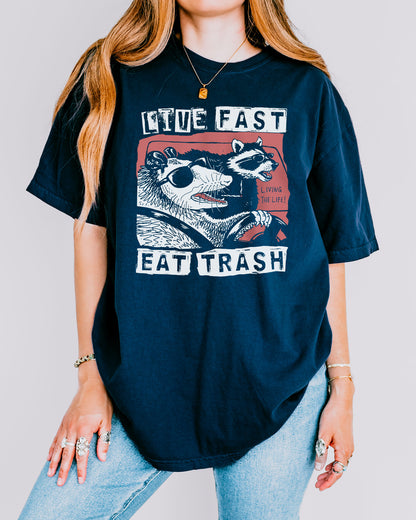 Woman wearing a true-navy-colored "Live Fast Eat Trash" shirt with a retro graphic of a raccoon and opossum driving a car.