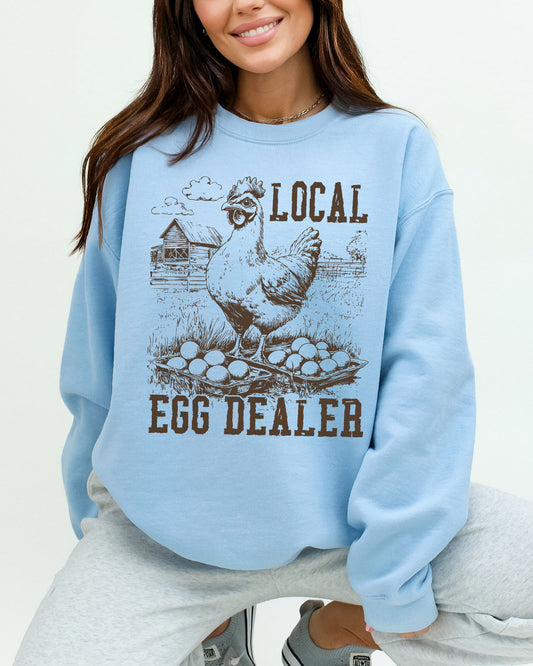 Woman wearing a light blue hoodie featuring a funny retro-style chicken on a farm graphic and the quote 'Local Egg Dealer'.