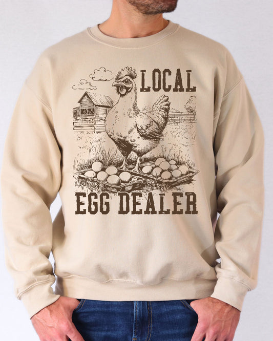 Man wearing a sand beige hoodie featuring a funny retro-style chicken on a farm graphic and the quote 'Local Egg Dealer'.