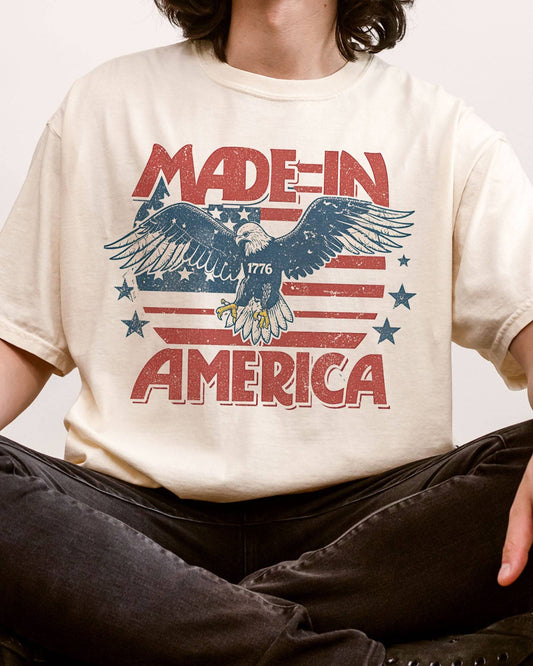 Close-up of man wearing "Made In America 1776" shirt in ivory color with retro graphic of a hawk in USA red-white-and-blue colors.