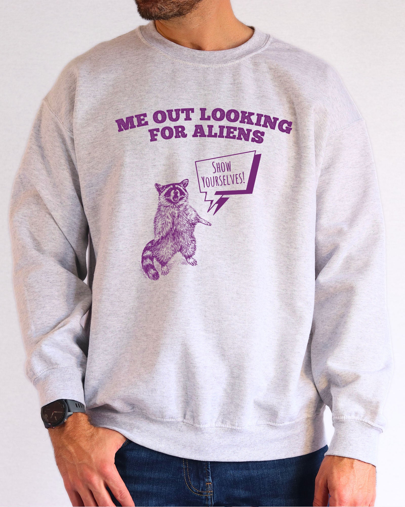 Model wearing ash color sweatshirt with 'Me Out Looking For Aliens' funny raccoon graphic design