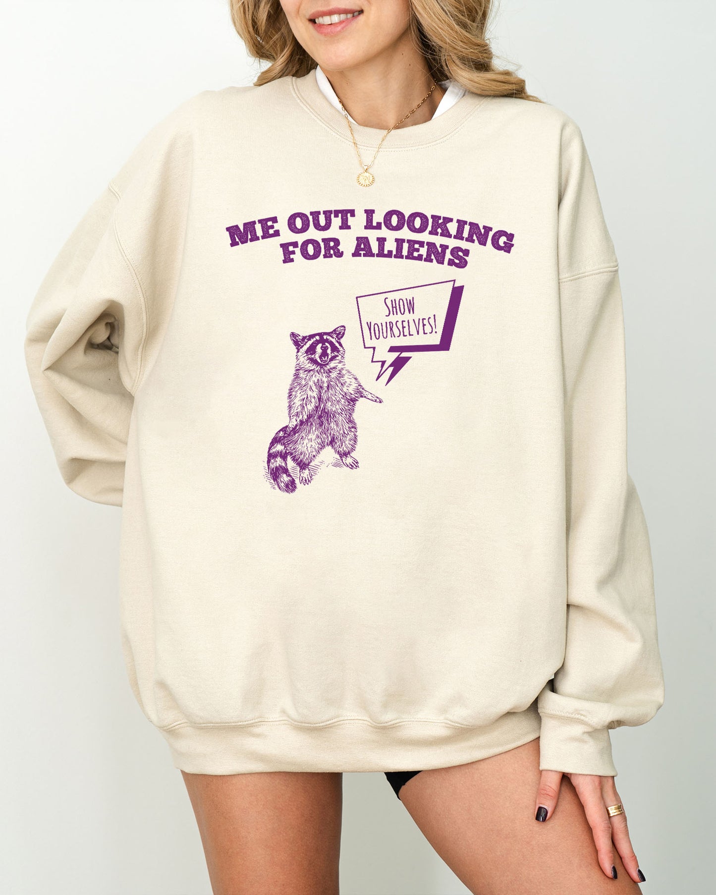 Model wearing sand color sweatshirt with 'Me Out Looking For Aliens' funny raccoon graphic design