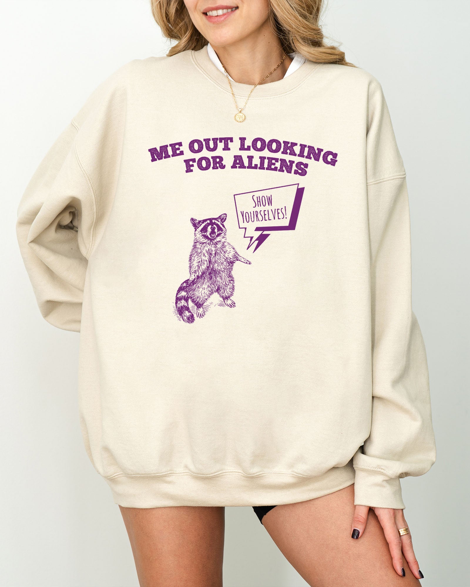Model wearing sand color sweatshirt with 'Me Out Looking For Aliens' funny raccoon graphic design