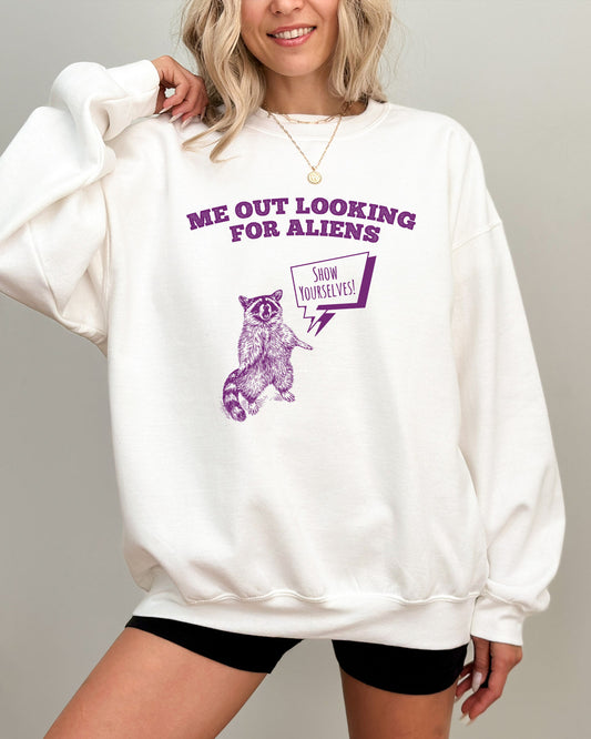 Model wearing white color sweatshirt with 'Me Out Looking For Aliens' funny raccoon graphic design
