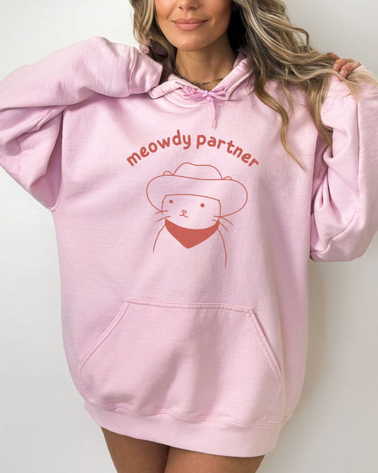 Woman wearing a light pink hoodie featuring a funny retro cowboy cat line-art graphic with the quote 'Meowdy Partner'.