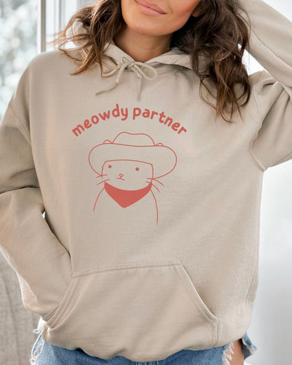 Woman wearing a sand beige hoodie featuring a funny retro cowboy cat line-art graphic with the quote 'Meowdy Partner'.