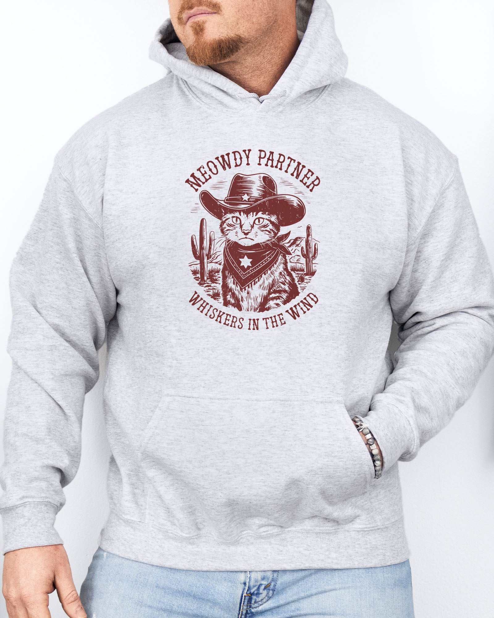 Man wearing an ash grayhoodie with a funny retro cowboy cat graphic and the text 'Meowdy Partner, Whiskers in the Wind' in a western-style design.