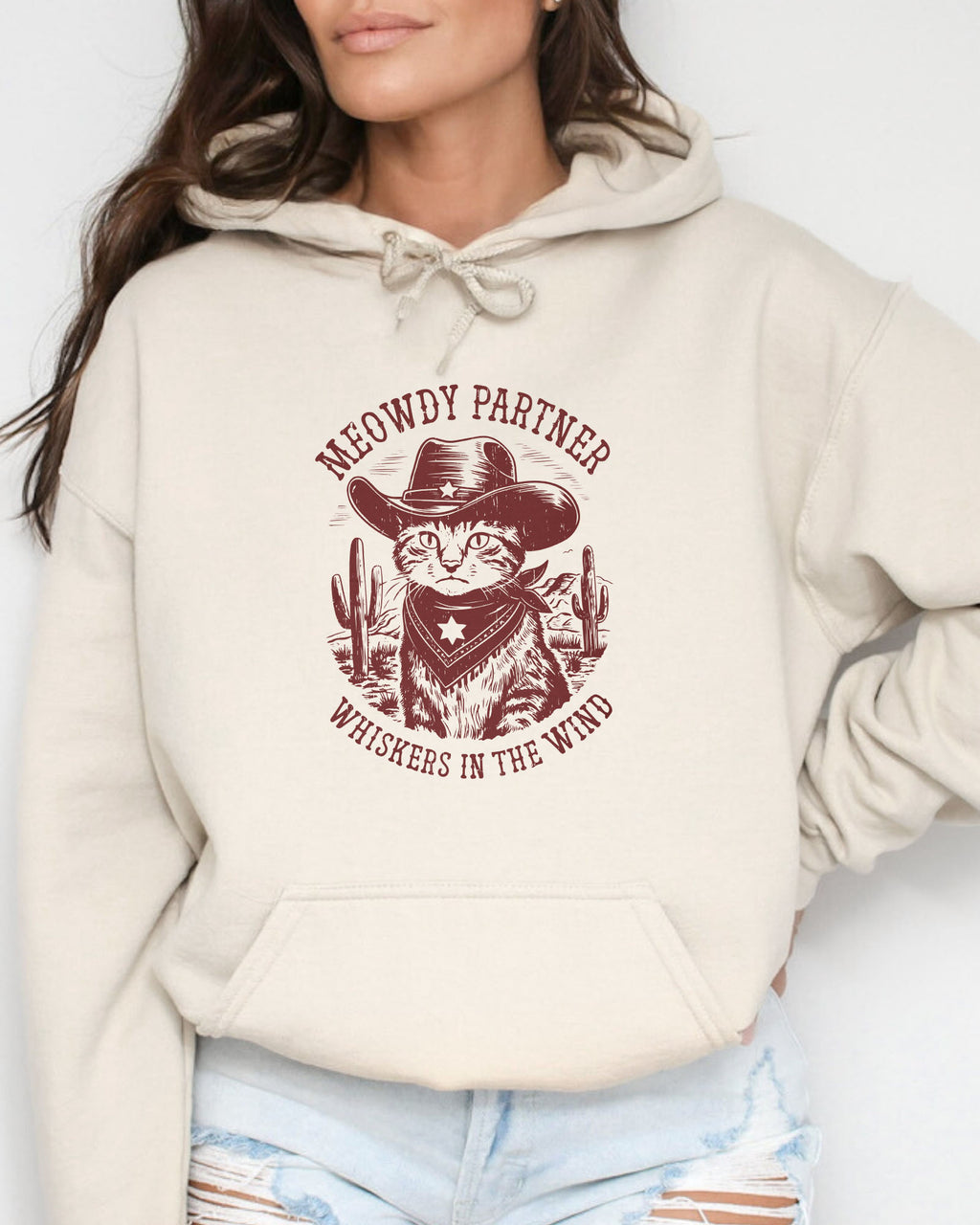 Woman wearing a sand beige hoodie with a funny retro cowboy cat graphic and the text 'Meowdy Partner, Whiskers in the Wind' in a western-style design.