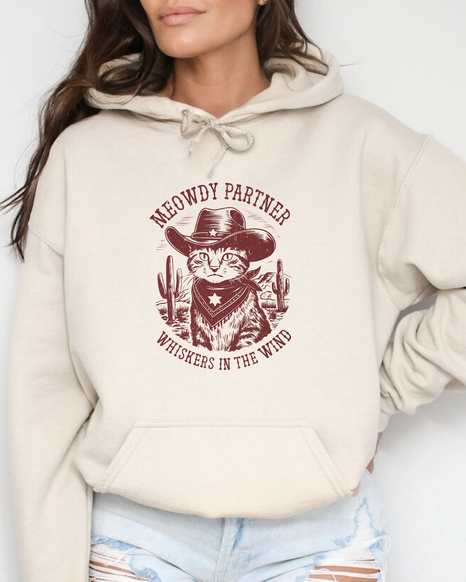 Woman wearing a sand beige hoodie with a funny retro cowboy cat graphic and the text 'Meowdy Partner, Whiskers in the Wind' in a western-style design.
