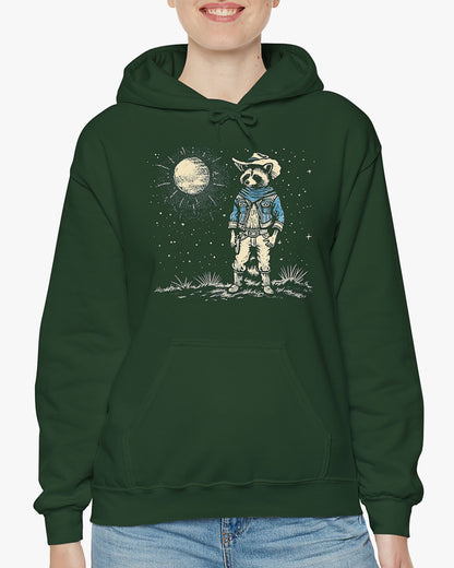Woman wearing a forest green hoodie featuring a retro Wild West graphic of a cowboy raccoon standing under a moonlit desert sky. The vintage-style design pops against the dark fabric.