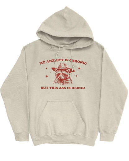 Flat lay of a sand beige 'My Anxiety Is Chronic But This Ass Is Iconic' hoodie featuring a funny retro cowboy raccoon graphic.