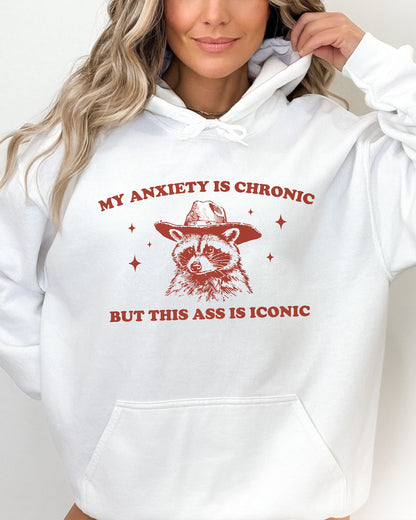 Woman wearing a white 'My Anxiety Is Chronic But This Ass Is Iconic' hoodie featuring a funny retro cowboy raccoon graphic.