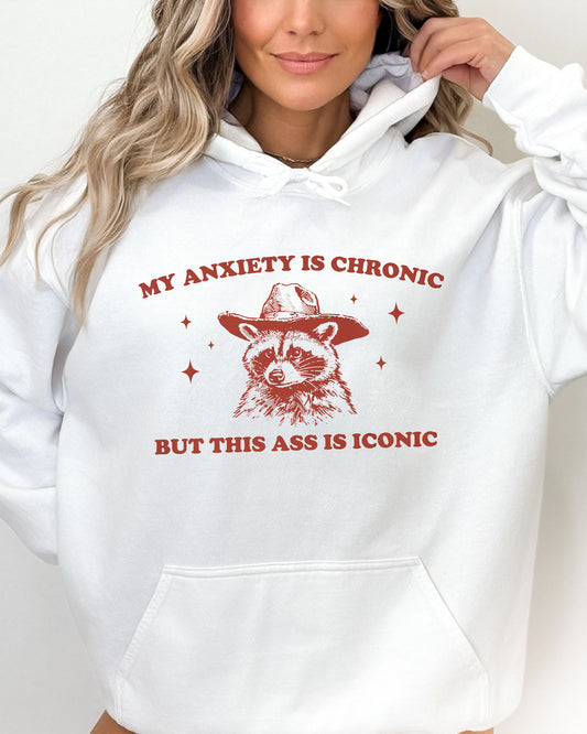 Woman wearing a white 'My Anxiety Is Chronic But This Ass Is Iconic' hoodie featuring a funny retro cowboy raccoon graphic.