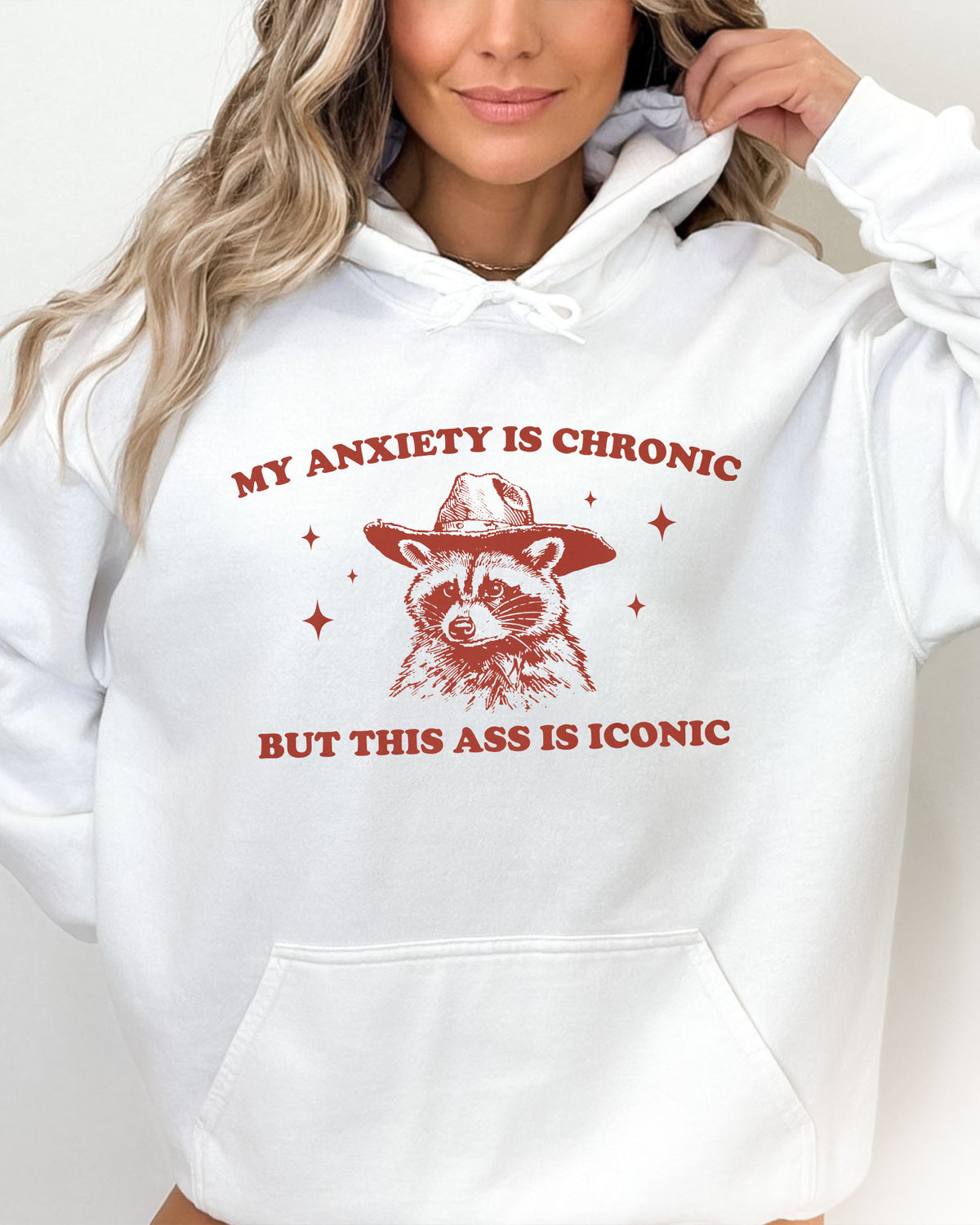 Woman wearing a white 'My Anxiety Is Chronic But This Ass Is Iconic' hoodie featuring a funny retro cowboy raccoon graphic.