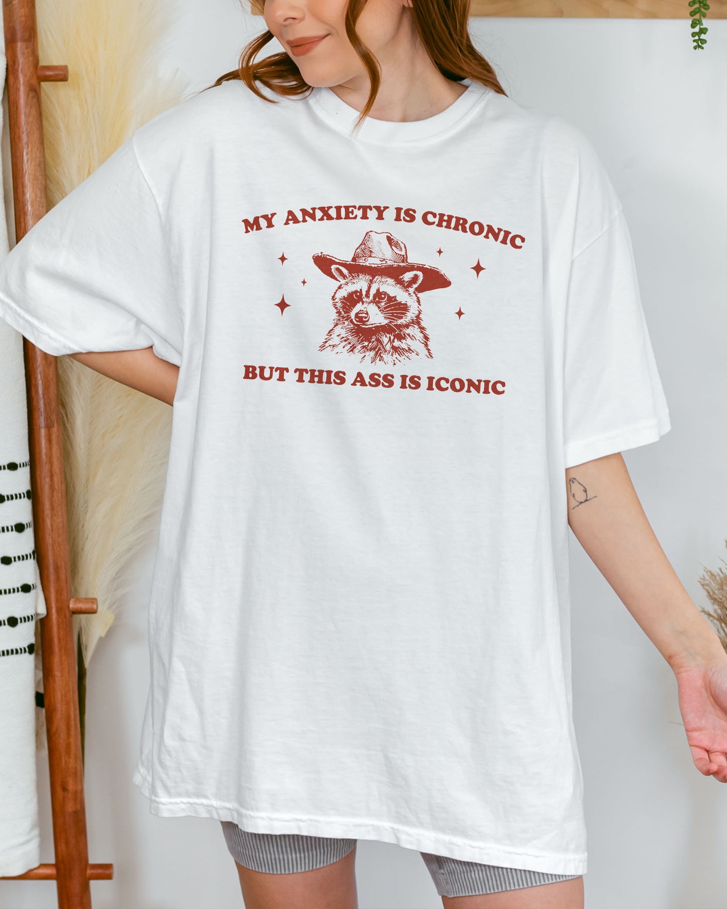 Woman wearing a white "My Anxiety Is Chronic But This Ass Is Iconic" shirt with a retro graphic of a cowboy raccoon.