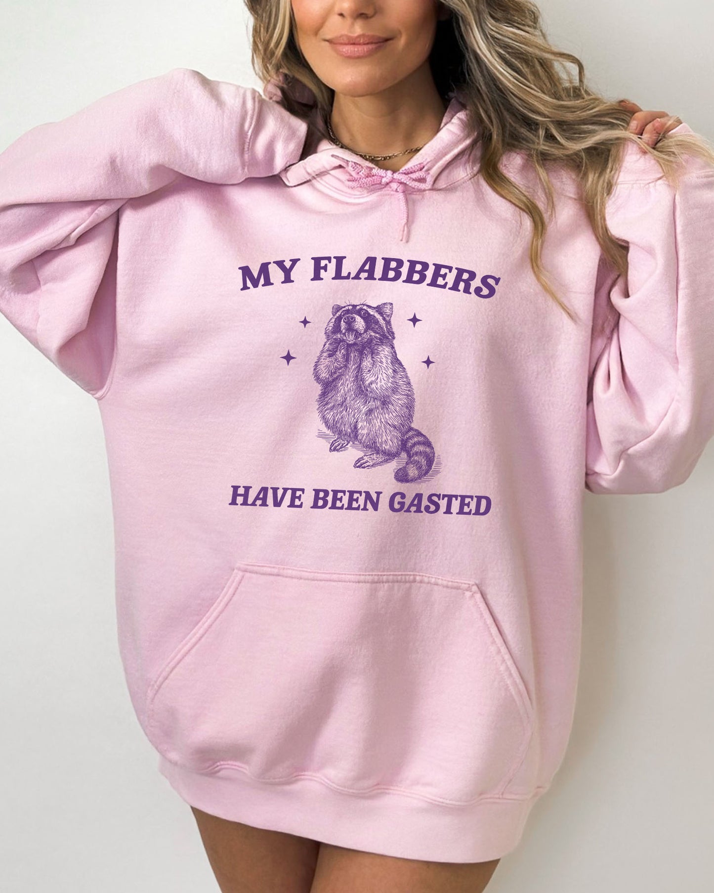Woman wearing a light pink 'My Flabbers Have Been Gasted' hoodie featuring a funny retro raccoon freaking out graphic.