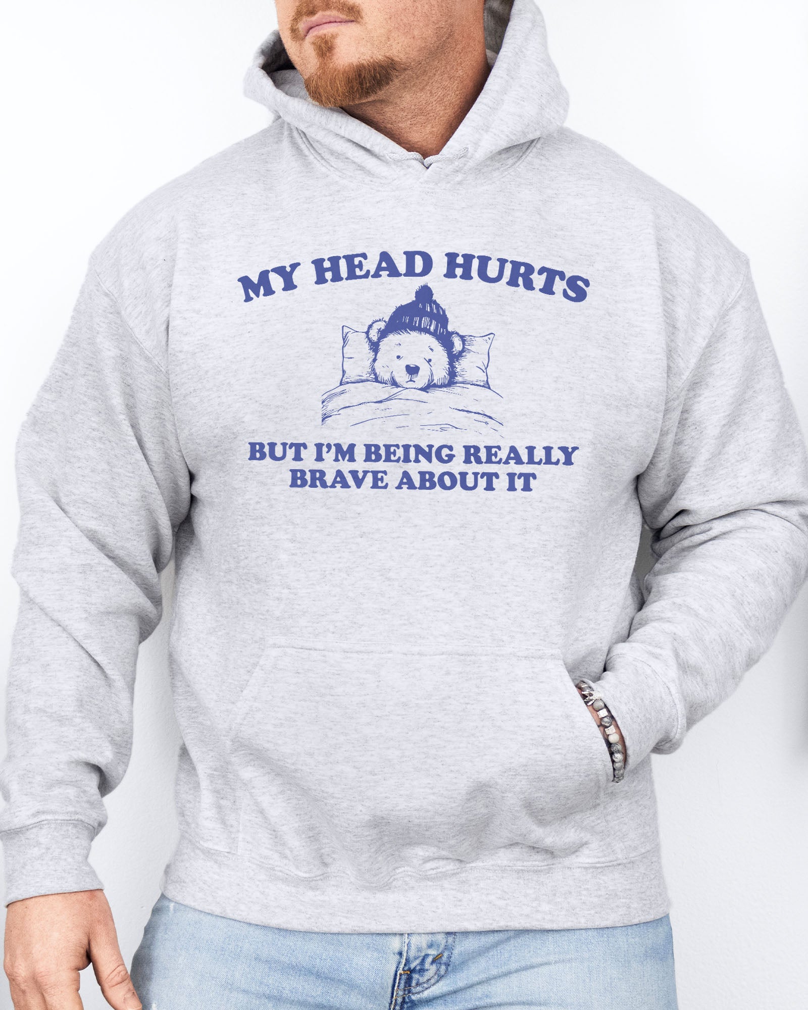 Man wearing an ash gray hoodie featuring a funny retro-style bear lying in bed with the quote 'My Head Hurts But I'm Being Really Brave About It'.