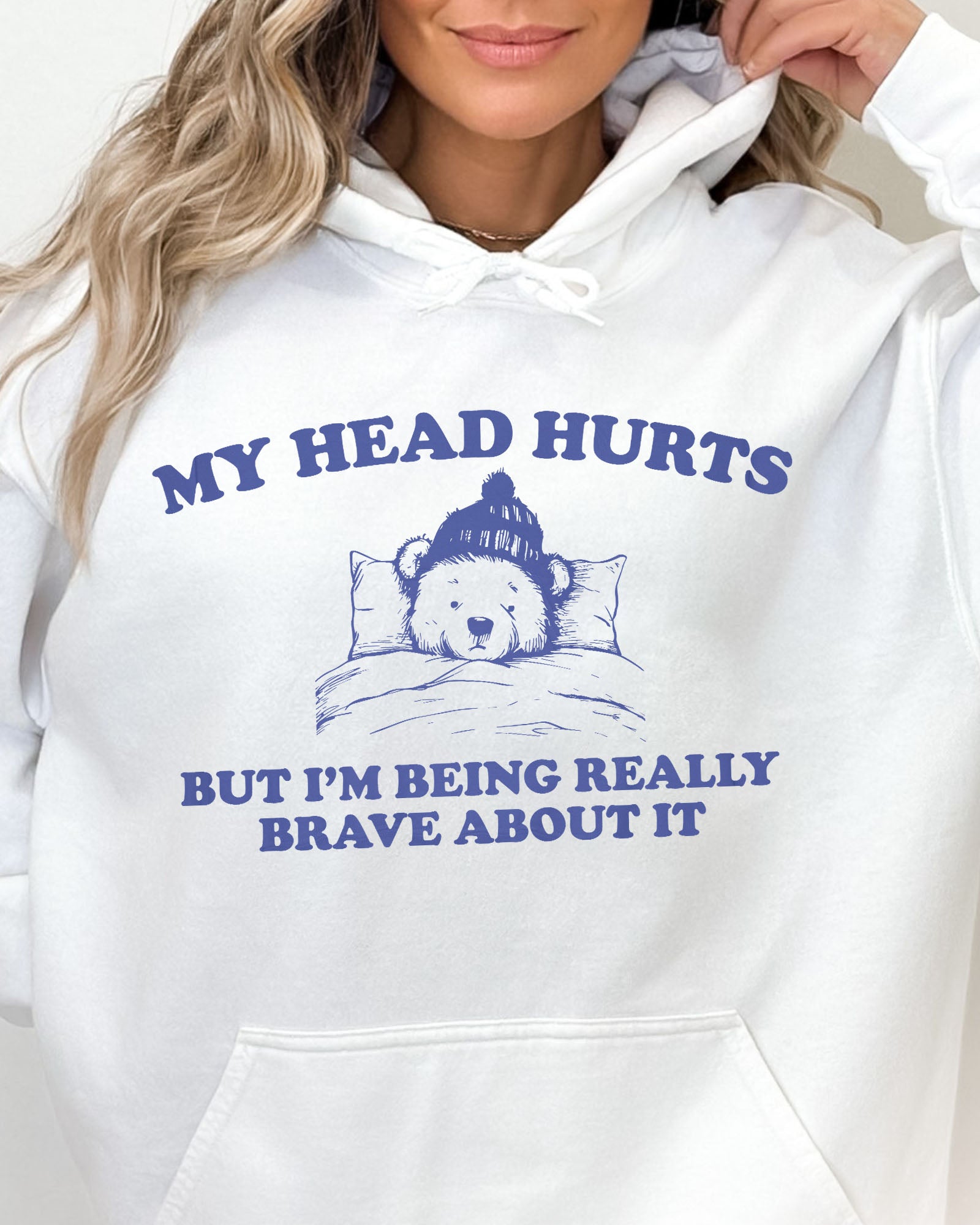 Close-up of a white hoodie featuring a funny retro-style bear lying in bed with the quote 'My Head Hurts But I'm Being Really Brave About It'.