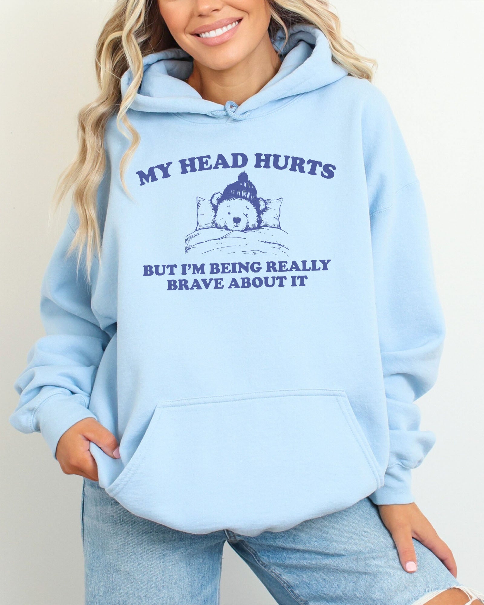 Woman wearing a light blue hoodie featuring a funny retro-style bear lying in bed with the quote 'My Head Hurts But I'm Being Really Brave About It'.