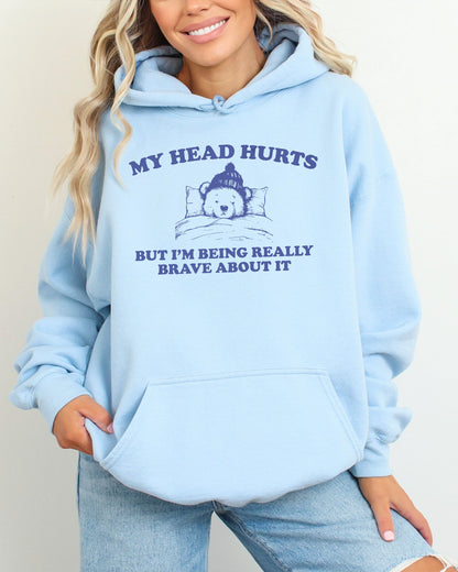 Woman wearing a light blue hoodie featuring a funny retro-style bear lying in bed with the quote 'My Head Hurts But I'm Being Really Brave About It'.