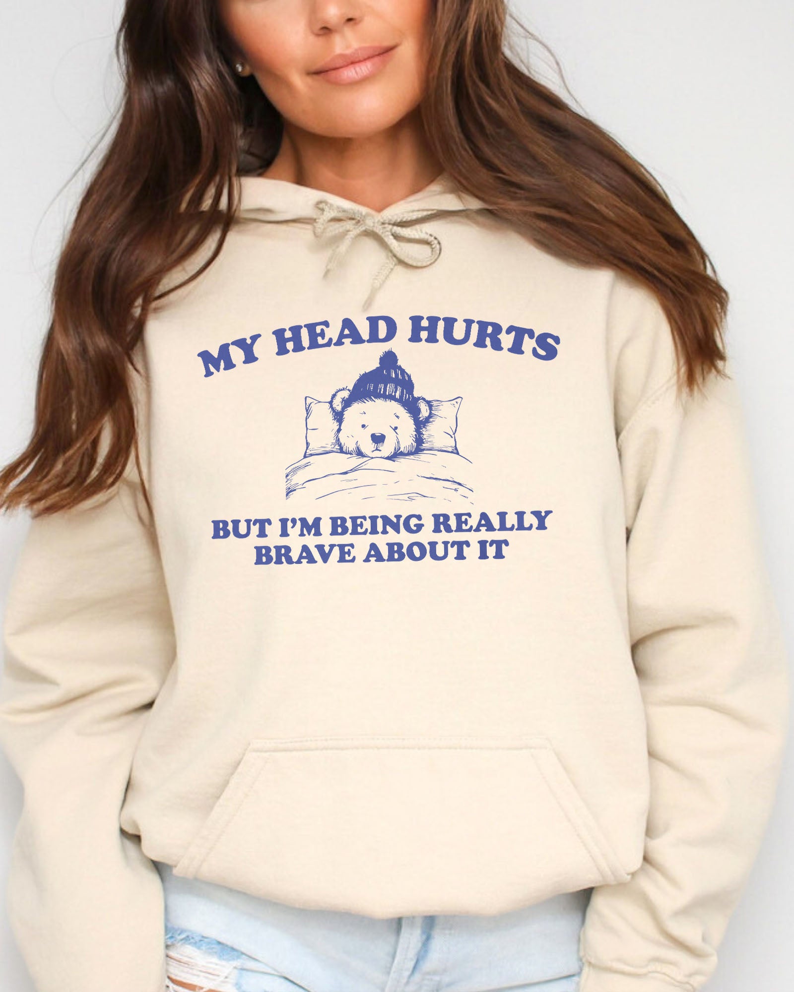 Woman wearing a sand beige hoodie featuring a funny retro-style bear lying in bed with the quote 'My Head Hurts But I'm Being Really Brave About It'.