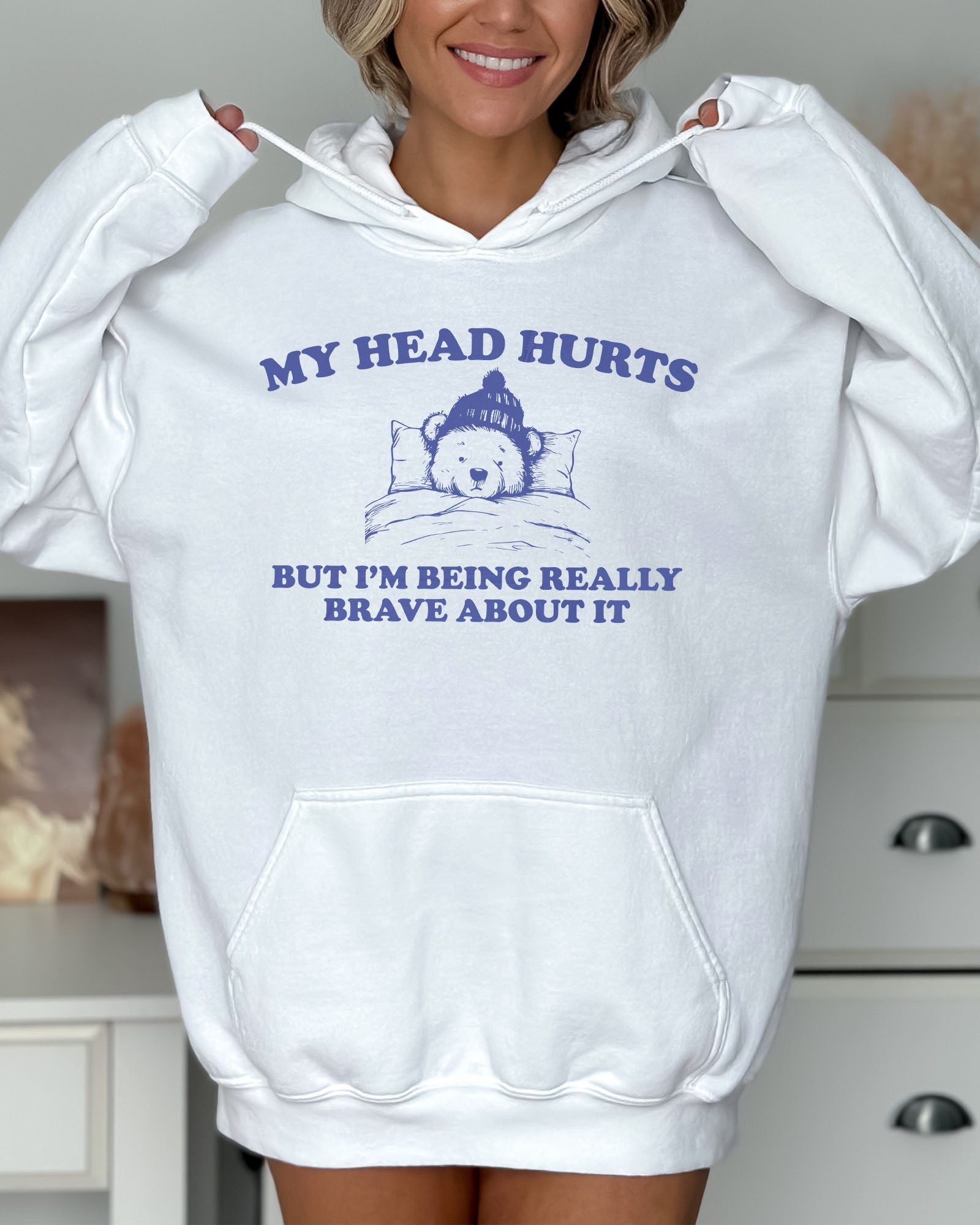 Woman wearing a white hoodie featuring a funny retro-style bear lying in bed with the quote 'My Head Hurts But I'm Being Really Brave About It'.