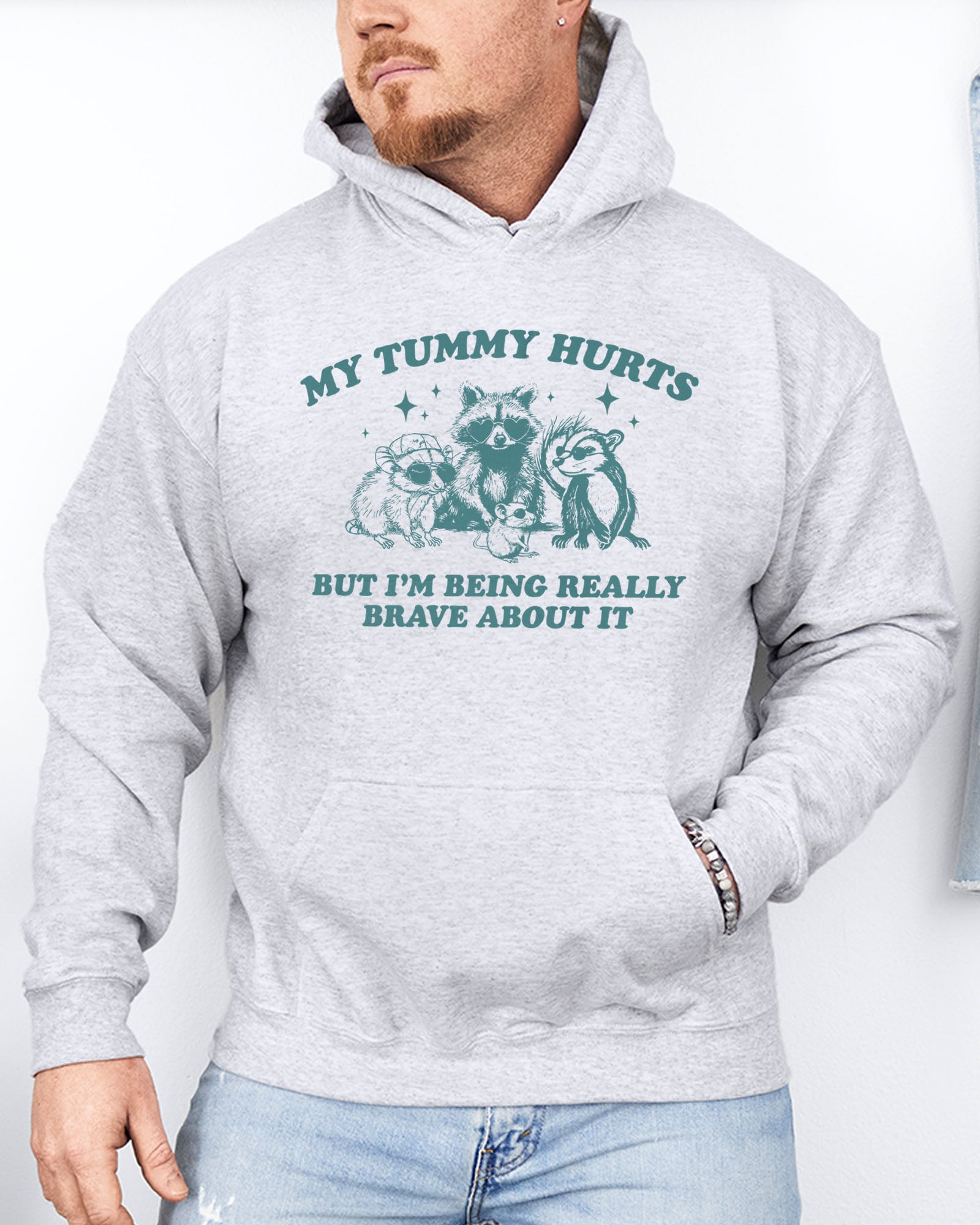 Man wearing an ash gray hoodie with a retro raccoon gang graphic and the quote 'My Tummy Hurts But I'm Being Really Brave About It'.